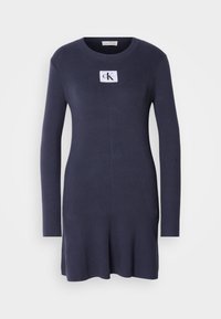 Marineblauw ribgebreid jurkje met lange mouwen en een uitlopende zoom. Heeft een witte patch met het logo "CK" op de borst. Zachte textuur.