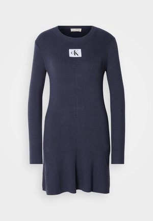 Marineblauw ribgebreid jurkje met lange mouwen en een uitlopende zoom. Heeft een witte patch met het logo "CK" op de borst. Zachte textuur.