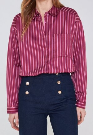 Femme portant une chemise à manches longues rayée violette rentrée dans un pantalon bleu foncé taille haute avec des boutons dorés.