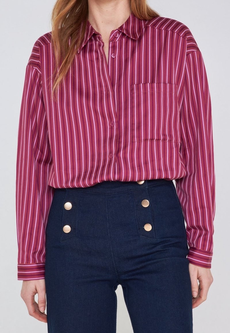 Femme portant une chemise à manches longues rayée violette rentrée dans un pantalon bleu foncé taille haute avec des boutons dorés.