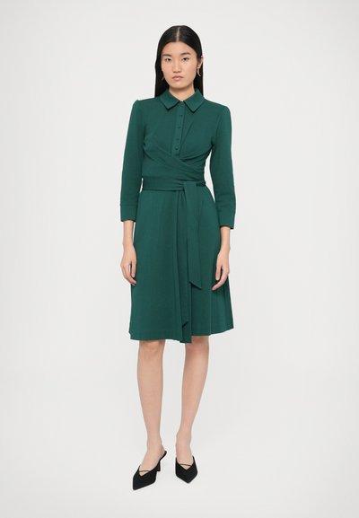 Πράσινο φόρεμα τύπου wrap με γιακά, κουμπιά μπροστά και μανίκια τριών τετάρτων. Διαθέτει ζώνη στη μέση που δένει και φουσκωτή φούστα που τελειώνει πάνω από τα γόνατα.