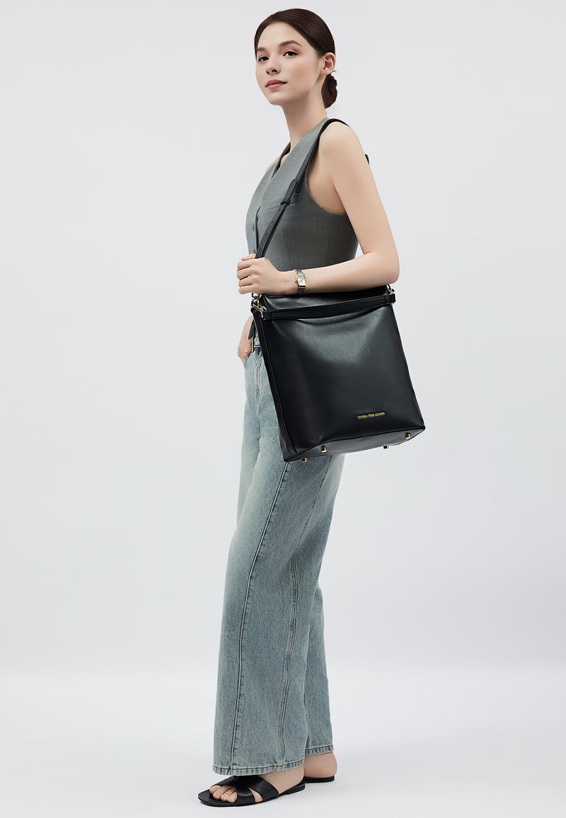Borsa a tracolla nera in pelle dal design minimalista, con dettagli dorati e una forma strutturata, portata da una persona in denim chiaro e sandali.
