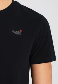 Černé bavlněné tričko s krátkým rukávem, které má na levé straně hrudi malou vyšívanou nášivku "SuperDry" v bílé a červené barvě.