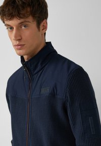 Veste en polaire bleu marine avec une partie inférieure texturée, une partie supérieure lisse, une fermeture éclair et un patch logo sur la poitrine. Comprend un col montant.