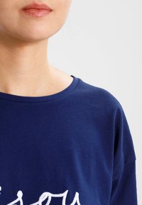 Detailní záběr osoby v námořnickém modrém mikině s bílým kurzívním textem, zobrazující krk, rty a ramena na světlém pozadí.