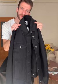 Schwarze Jeansjacke mit großem Kragen, Fronttaschen und silbernen Knopfdetails. Der Stoff wirkt robust mit einer leichten Textur.