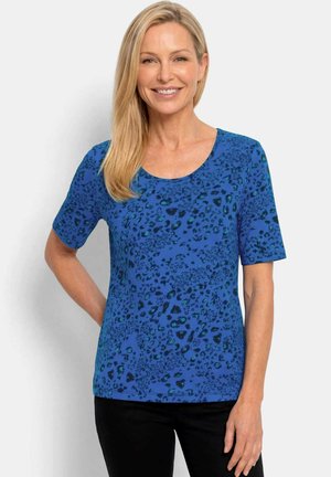 Frau mit langen blonden Haaren, die ein blaues kurzärmeliges Leopardenmuster-Shirt und schwarze Hose trägt, lächelnd mit der Hand in der Hüfte.