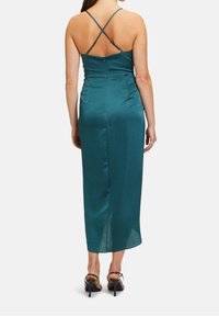 Robe midi en satin sarcelle avec des bretelles croisées dans le dos, taille cintrée et fente latérale subtile. Accentuée par une fermeture éclair et une texture lisse.