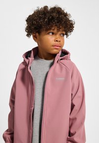 Weicher, rosa Reißverschlussjacke mit Kapuze, Seitentaschen und Logodetail. Getragen über einem hellgrauen Shirt, das eine glatte Textur zeigt.