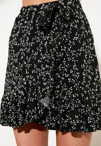Zwarte, geribbelde rok met een wit bloemmotief, met een rucheszoom en een knoopdetail in de taille. De stof lijkt lichtgewicht en gestructureerd.