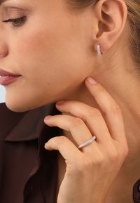 Silver hoop earrings with small sparkling accents, paired with a plain silver ring, on a hand near a face with neutral makeup, side profile.