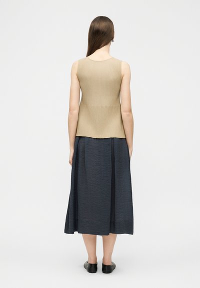 Femme aux longs cheveux bruns portant un haut côtelé sans manches beige, une jupe midi bleu foncé et des chaussures plates noires, debout de dos.