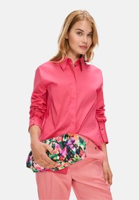 Bright pink long-sleeve button-up shirt paired with striped pink pants. Holding a floral-patterned clutch bag featuring vibrant colors.