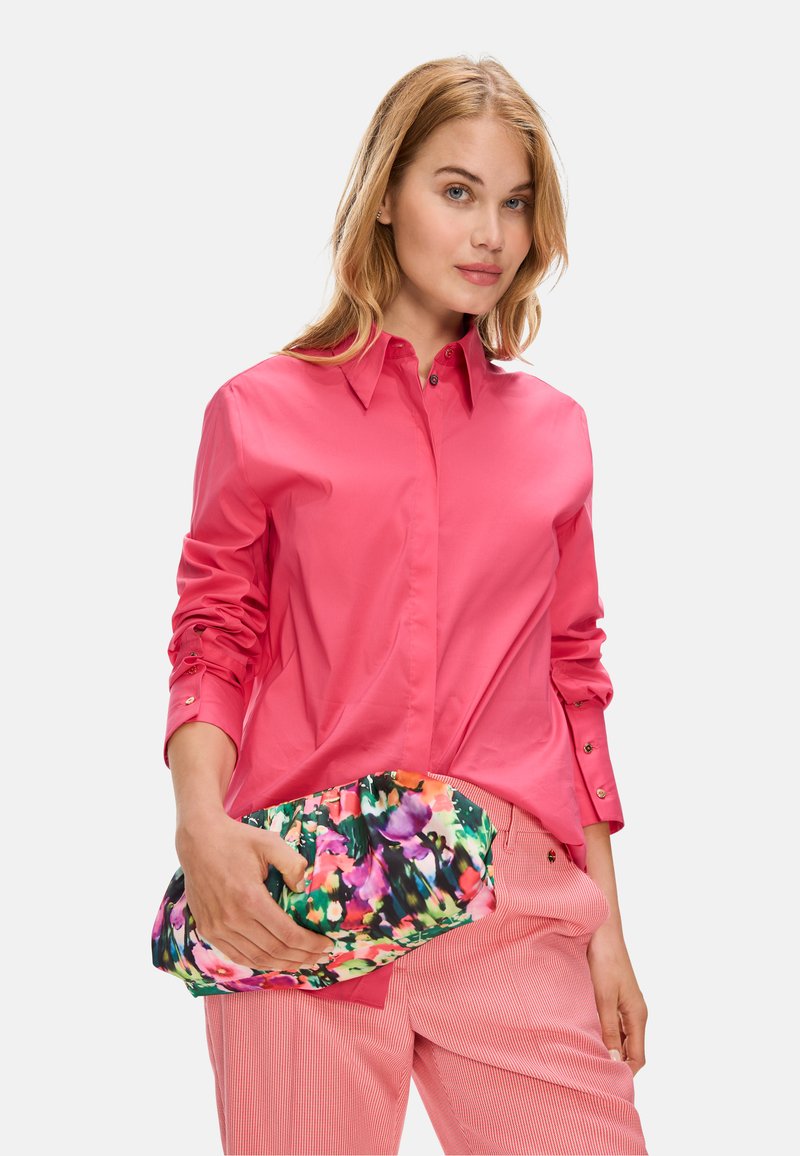 Bright pink long-sleeve button-up shirt paired with striped pink pants. Holding a floral-patterned clutch bag featuring vibrant colors.
