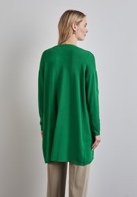 Haut en maille vert à manches longues avec une coupe décontractée, présentant des fentes sur les côtés et un ourlet arrondi. Associé à un pantalon large beige.