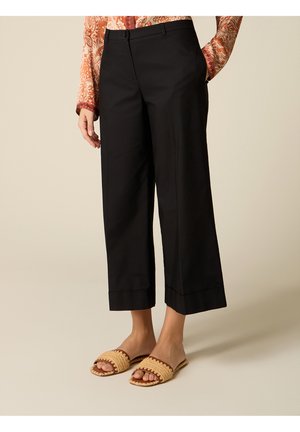 Donna che indossa pantaloni neri larghi a gamba corta, camicetta a maniche lunghe con motivo, e sandali beige intrecciati senza lacci, in piedi su uno sfondo semplice.