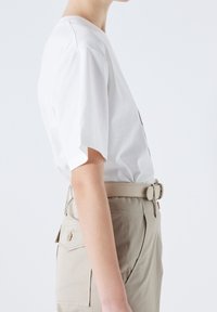 Une personne portant une chemise blanche à manches courtes rentrée dans un pantalon beige taille haute avec une ceinture assortie et une poche à rabat boutonnée.