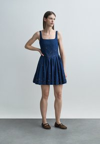 Vrouw staand met één hand op haar heup, gekleed in een mouwloze blauwe bloemenjurk en luipaardprint ballerina's tegen een effen achtergrond.