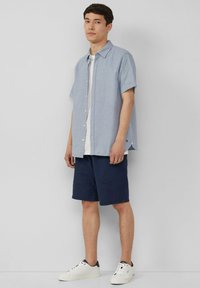 Kurzärmliges Hemd mit Knöpfen aus hellblauem und weißem gestreiftem Stoff, kombiniert mit marineblauen Shorts und weißen Sneakern mit dunklen Akzenten.