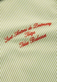Broderie cursive rouge disant « Sant Antoni de Portmany Ibiza Îles Baléares » sur un tissu rayé diagonal vert et blanc.