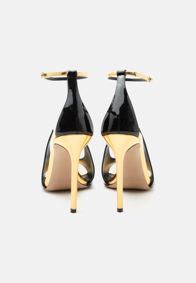 Μαύρες ψηλές γόβες από πατέντα δέρμα με λεπτά χρυσά τακούνια στιλέτο και χρυσά λουριά στον αστράγαλο, σε προβολή από πίσω σε λευκό φόντο.