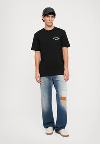 T-shirt nera in cotone con logo bianco, abbinata a jeans in denim blu chiaro con toppe beige e marroni, indossando scarpe bianche e un berretto.