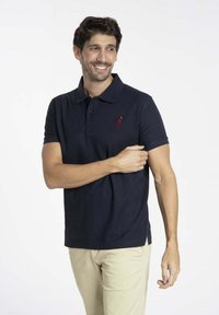 Polo shirt bleu marine avec un col, des manches courtes et un logo brodé bordeaux sur la poitrine. Fabriqué en tissu en coton lisse.