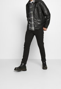Schwarze Lederjacke mit Taschen, Karottenhemd in Grün- und Weißtönen, schwarze Slim-Fit-Hose und schwarze Stiefeletten mit strukturiertem Sohlen.