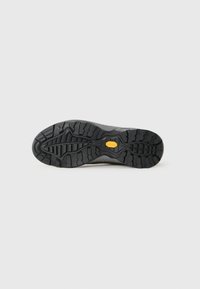Semelle extérieure en caoutchouc noir avec un motif de crampons profonds et un logo jaune distinctif. Le design présente une surface texturée pour une meilleure traction et durabilité.