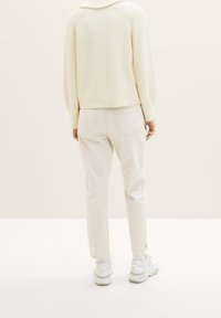 Personne portant un pull écru, un pantalon beige clair avec des poches arrière, et des baskets blanches, debout devant un fond beige uni.