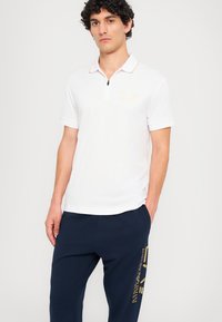 Balta polo marškinėliai su trumpu užtrauktuku, auksiniais akcentais ir EA7 logotipu, derinami su tamsiai mėlynomis treningo kelnėmis, kurių šone yra auksinė užrašai.