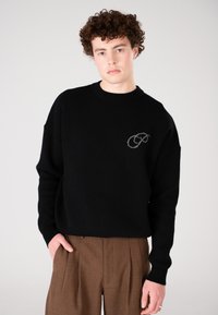 Junger Mann mit lockigem Haar, der einen schwarzen Pullover mit einem kleinen grauen Logo und braunen bügelgefalteten Hosen trägt, und vor einem schlichten Hintergrund steht.