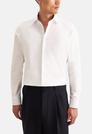 Chemise blanche à manches longues avec un col boutonné, en tissu texturé, et un détail de col intérieur contrastant, associée à un pantalon noir.
