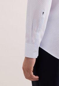 Wit overhemd met lange mouwen en een lichtblauw geometrisch patroon; voorzien van manchetten met knoopsluiting en een kleine geborduurde bloem op de mouw.