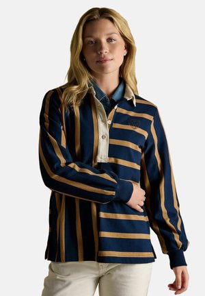 Young woman with blonde hair wearing a navy and tan striped rugby shirt, light-colored pants, and pearl earrings against a plain background.