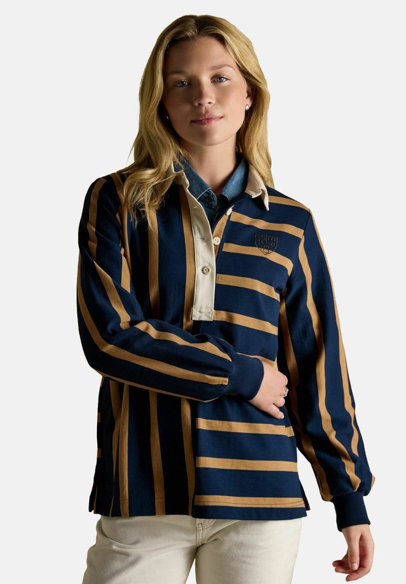Young woman with blonde hair wearing a navy and tan striped rugby shirt, light-colored pants, and pearl earrings against a plain background.