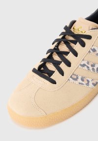 Baskets en suède beige avec lacets noirs, présentant un motif léopard sur le côté. Semelle en caoutchouc avec une finition texturée. Accent logo doré.
