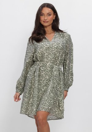 Femme portant une robe à manches longues, longueur genoux, avec un petit motif floral vert et blanc, debout devant un fond blanc uni.