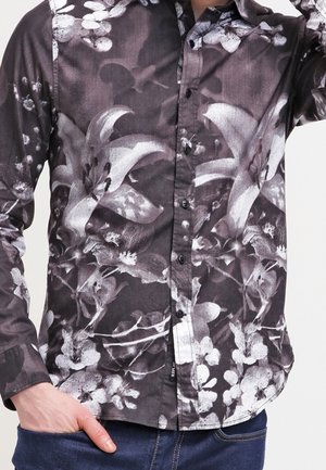 Man wearing a dark long-sleeve button-up shirt with a large white floral print and blue jeans, hand in pocket.