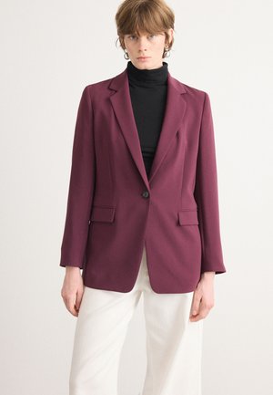 Maroon blazer with a single button and two front pockets, made of sleek fabric. Worn over a black turtleneck with white trousers.