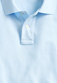Polo bleu clair avec col et deux boutons blancs sur la patte de boutonnage, montré en gros plan.