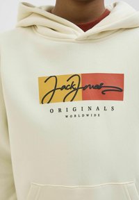 Kremo spalvos džemperis su priekine kišene, su geltonai raudonu stačiakampiu logotipu. Tekstas: „Jack Jones ORIGINALS WORLDWIDE“ juoda spalva.