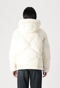 Balta pūkaina jaka ar kapuci, ar kvadrātveida dizainu un dimanta formas dūrienu. Gluda tekstūra, brīva piegriezuma un saīsināts garums.