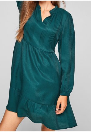 Vrouw die een teal langarmige jurk draagt met geplisseerd lijfje, golvende halslijn en gerafelde zoom, poserend tegen een effen grijze achtergrond.