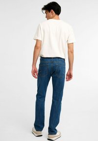 Blå denimjeans med rak passform, synliga sömmar och två bakfickor, tillsammans med en enkel vit T-shirt och lätta skor.