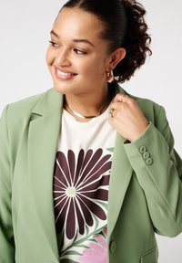 Groene op maat gemaakte blazer met een gladde textuur, gecombineerd met een crèmekleurig shirt met een groot donkerpaars en pastel bloemenpatroon. Gouden sieraden als accenten.