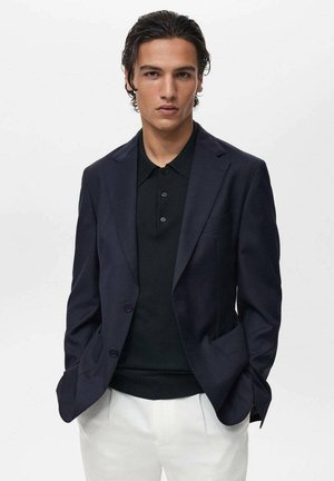 Navy blazer with notched lapels, worn over a black polo shirt. White trousers. Smooth fabric, classic design, tailored fit.