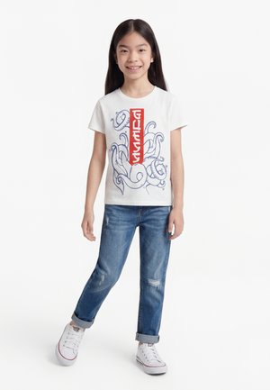 Χαμογελαστή κοπέλα που φοράει λευκό T-shirt με κόκκινο κατακόρυφο λογότυπο και σχέδιο με μπλε πλοκάμια οκτώποδα, μπλε τζιν με ρεβέρ και λευκά αθλητικά παπούτσια.