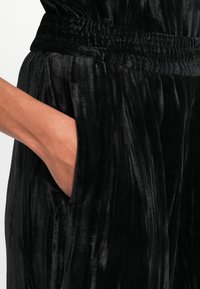 Combinaison en velours noir avec texture verticale, dotée d'une taille élastique et de poches latérales. Gros plan sur une main dans la poche, mettant en avant le tissu.