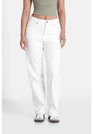 Kvinde iført hvide højttaljede jeans, grøn crop top, sneakers og et armbånd, stående på en lys baggrund.